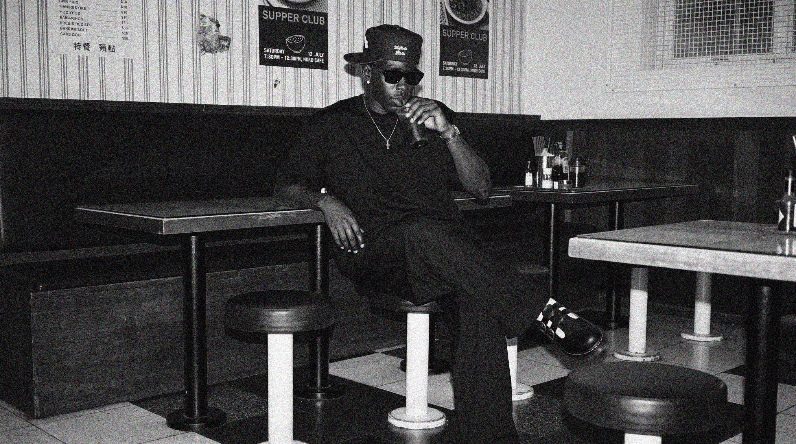 A black and white photograph of a man sitting in a diner booth, wearing a baseball cap, sunglasses, and a dark outfit. The scene includes retro diner tables, stools, and wall posters.