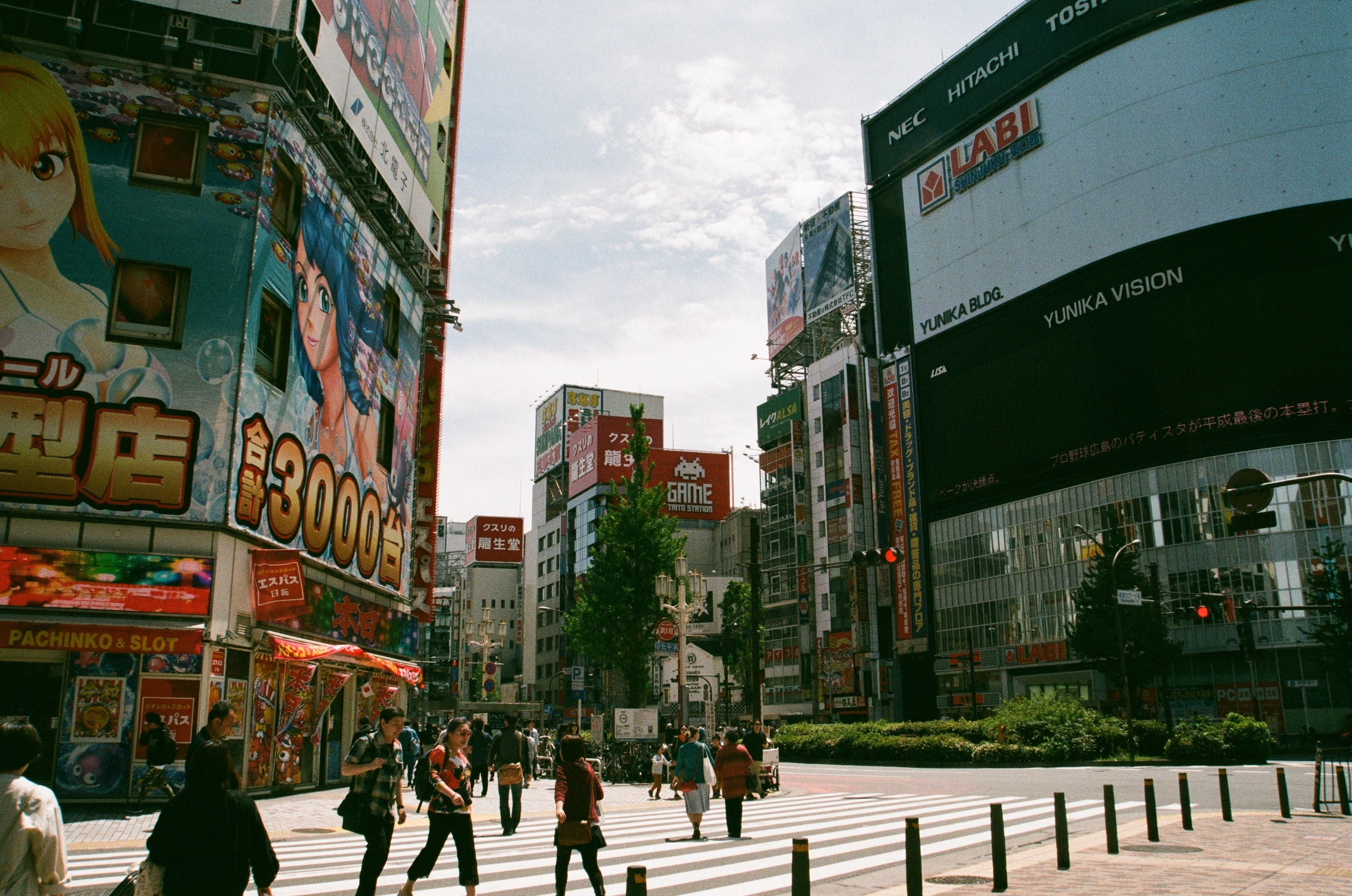 A wide street scene in a Japanese city with pedestrians crossing a crosswalk, surrounded by tall commercial buildings, colorful storefronts, and large billboards under a bright sky.